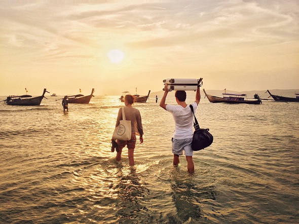 Young couple at Ao Nang beach in Krabi province walking into the ocean to long tail boats during low tide and holding their bags over their heads.