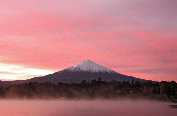 Chile's snow-capped Villarrica Volcano stands against a pink sky as mist rises from the lake in the foreground.