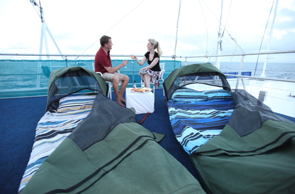 A couple share a toast on the top deck of Reefworld pontoon in the Whitsundays, with their swags in the foreground.