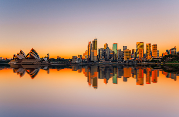 The Sydney Opera House and city skyline reflect in the harbour at sunset.