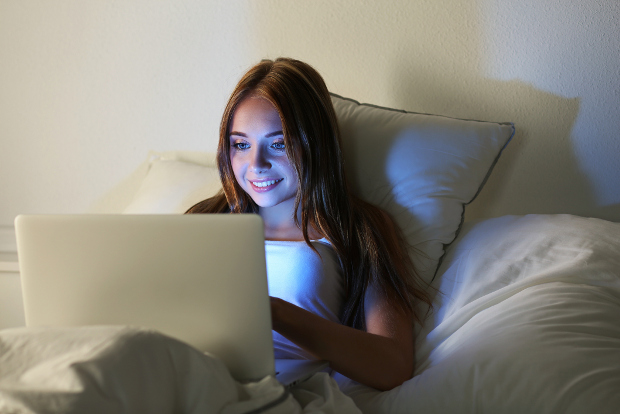 A young woman watches movies on her laptop in bed.