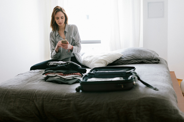 A woman sitting on a bed looking at her phone while packing her suitcase
