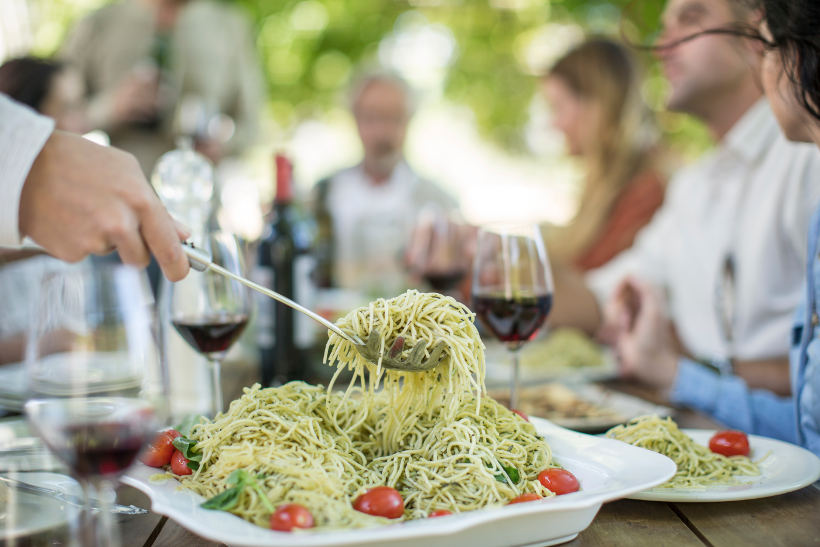 table of people with red wine glasses and someone serving pesto pasta