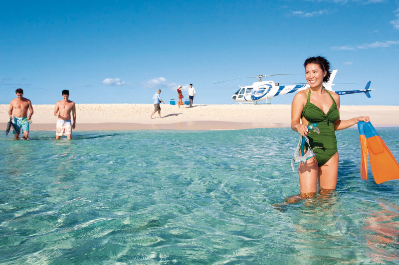 Woman holds snorkel gear in shallow water with helicopter and others behind her