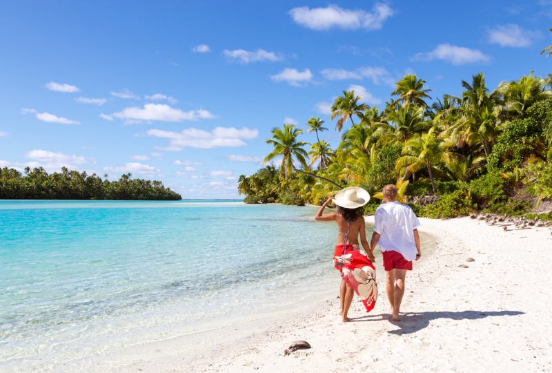 A couple walks across a white sandy beach among the coconut palms with a forested islet visible over an azure lagoon.