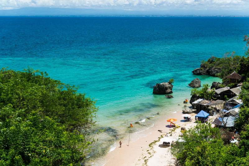 A view looking down on Bali's Padang Padang Beach, featuring umbrellas, huts, hillside, white sand and blue sea.