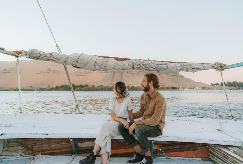 two people sit on the edge of a felucca with the sail and bank of the nile in background. one presents masc and has a brown shirt and jeans with long tied up hair, the other presents femme and wears white cotton top and pants with a scarf tied in hair  