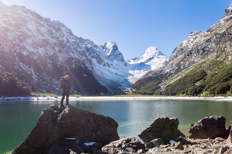 man standing on rock infront of lake and snow capped mountains