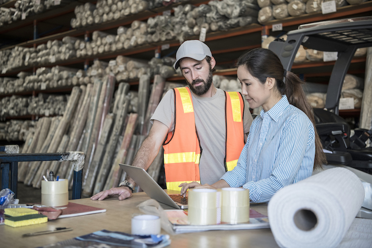 Woman using laptop in warehouse with male employee watching