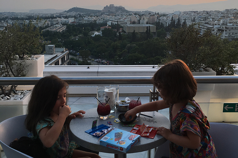 Two little girls sit on a hotel balcony with the Acropolis in the background.