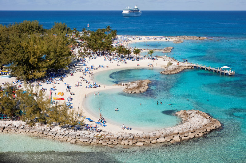 Passengers relax on a beach in the Caribbean, with a cruise ship in the distance.