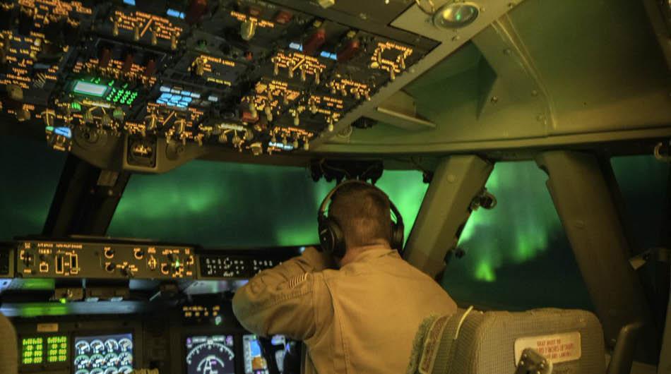 A male presenting pilot in cockpit of a plane with many lit up buttons, dials and levers. Green aurora can be seen through the windows