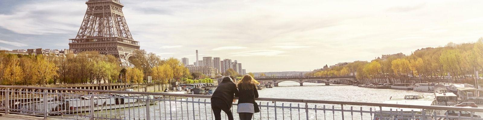Femme and masculine presenting people stand on bridge over looking Seine River in Paris. the Eiffell Tower is in the background.