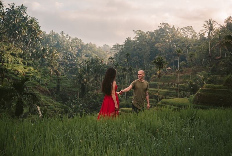 A femme presenting person wearing a bright red long dress holds the hand of a male presenting person wearing a green shirt. They are in a rice paddy field in Ubud, Bali. 