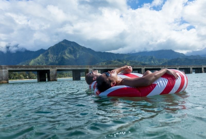 two male appearing people float on red and white striped floaties in the ocean with volcanic mountains of hawaii in backgroung 