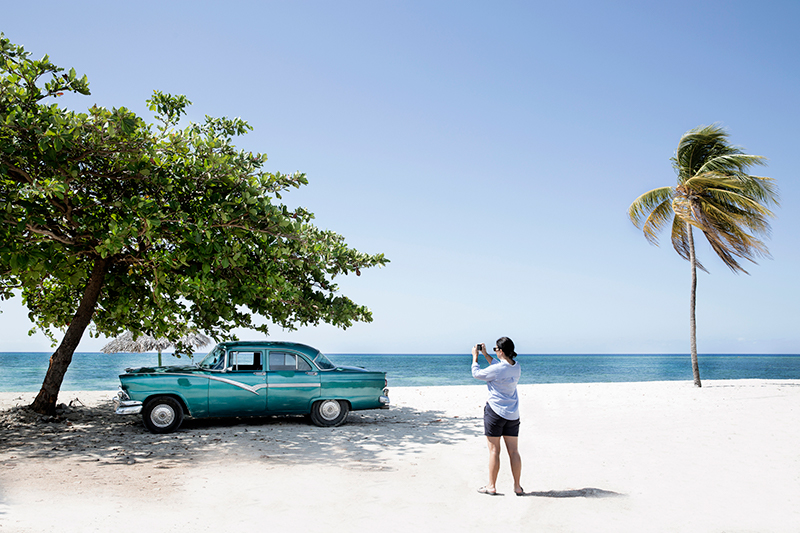 A woman takes a photograph of a vintage car on a beach in Cuba.
